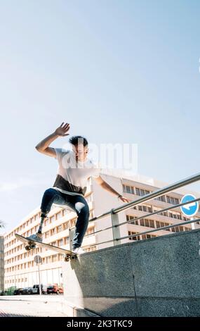 From below young guy with leg prosthesis jumping on skateboard above ground against modern building on sunny day Stock Photo