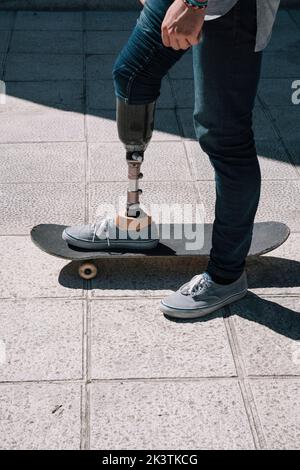 Side view of young guy with leg prosthesis standing and riding skateboard on city street Stock Photo