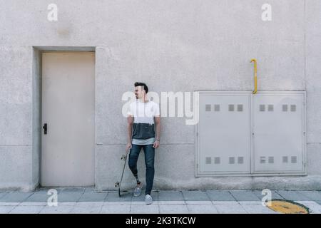 Young male with leg prosthesis smiling away while holding skateboard and standing against concrete building on street Stock Photo