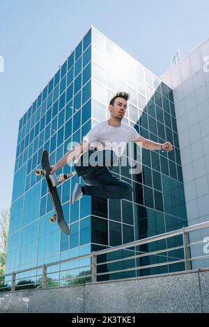 From below young guy with leg prosthesis jumping on skateboard above ground against modern building on sunny day Stock Photo