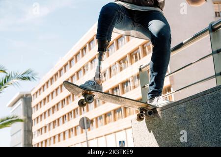 From below young guy with leg prosthesis jumping on skateboard above ground against modern building on sunny day Stock Photo