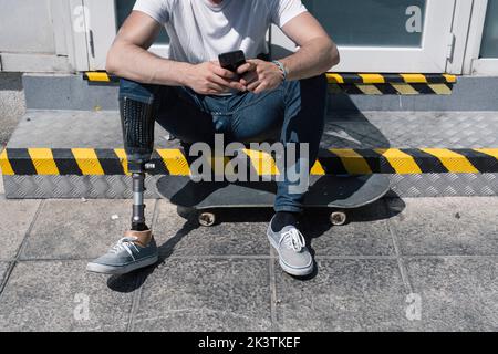From above anonymous male with artificial leg browsing smartphone while sitting on step near skateboard on sunny day on city street Stock Photo