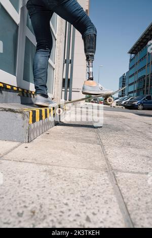 From below young guy with leg prosthesis jumping on skateboard above ground against modern building on sunny day Stock Photo