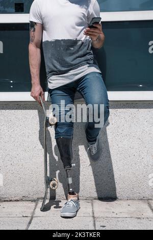 From above anonymous male with artificial leg browsing smartphone while standing holding skateboard on sunny day on city street Stock Photo