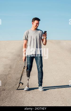 From above anonymous male with artificial leg browsing smartphone while standing holding skateboard on sunny day on city street Stock Photo