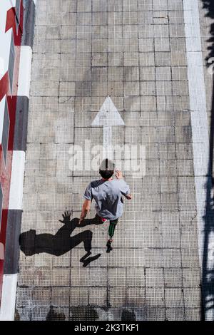 from above young guy with leg prosthesis running up paved slope against shabby building during workout on city street Stock Photo