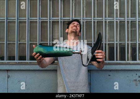 Young man smiling and demonstrating leg prosthesis while standing near building on city street Stock Photo