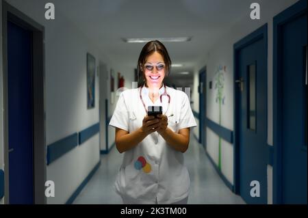 Glad adult woman in medical uniform smiling and browsing smartphone while walking along corridor in children hospital Stock Photo