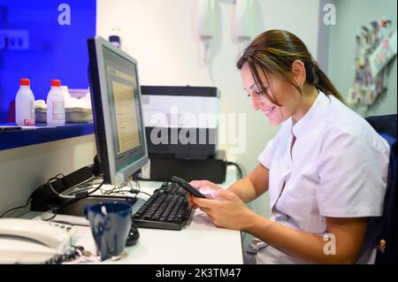 Side view of happy woman in medical uniform using computer while texting on mobile phone sitting at table in hospital lab Stock Photo