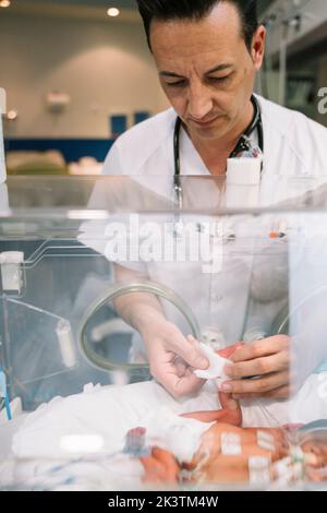 Doctor male attentively taking care of faceless newborn baby lying in incubator in clinic Stock Photo