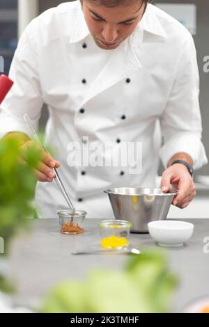 attentive man in white chef jacket preparing dish in stainless bowl and using metal tool at restaurant kitchen Stock Photo