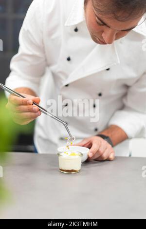 attentive man in white chef jacket preparing dish in glass bowl and using metal tool at restaurant kitchen Stock Photo