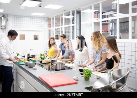 Group of multi ethnic people standing and learning by table while man demonstrating process of cutting vegetables in professional kitchen Stock Photo