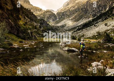 Side view of unrecognizable adventurer with backpack standing on stone amidst shallow river flowing through rocky Pyrenees mountains in summer day Stock Photo
