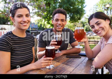 Medium-shot of three friends looking at camera while having a good time drinking beer at a trendy outdoors bar Stock Photo