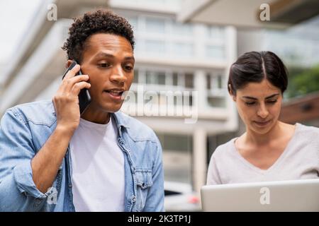 Mid-shot of African-American man speaking on mobile phone and Latin-American woman working on laptop computer outdoors Stock Photo
