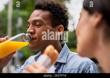 Side view shot of African-American man drinking orange juice and his Latin-American female friend eating a sandwich Stock Photo