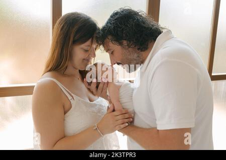 From above Hispanic parents embracing and admiring infant while sitting near pillows in bedroom at home Stock Photo