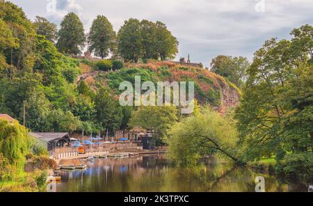 A tranquil scene where a tree arches over a river adjacent to moored boats. A hill is beyond with a monument on the skyline. A cloudy sky is above. Stock Photo