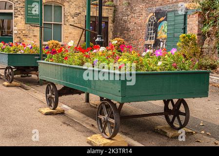 An old railway station trolley has been adapted as a flower bed with a colourful display of annual flowers. The wheels are on blocks of stone. Stock Photo