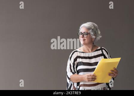 Middle aged Hispanic female in glasses holding folder with documents and looking away against gray background Stock Photo