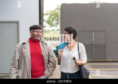 Positive Hispanic coworkers in stylish clothes talking and laughing while walking on sidewalk near building on city street in daytime Stock Photo