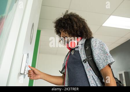 From below of side view of Hispanic man with long curly hair and headphones on neck in casual clothes standing and pressing button on security panel i Stock Photo