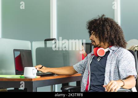 Side view of pensive Hispanic man with curly hair and headphones on neck sitting at wooden table and working on project on laptop in workspace in Cost Stock Photo