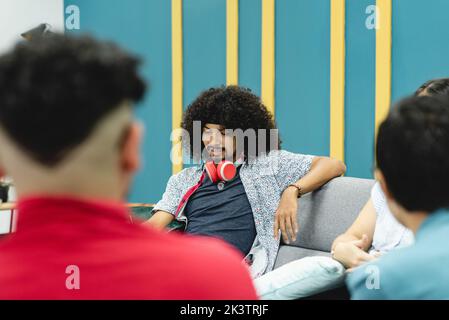 Calm Hispanic male with curly hair and headphones on neck in casual clothes sitting on comfortable sofa among unrecognizable people in modern workspac Stock Photo