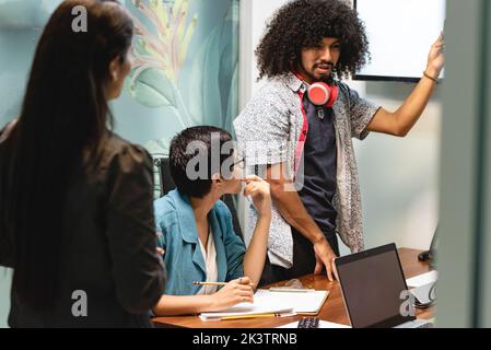 Pensive Hispanic man with curly hair in casual clothes standing and leaning on table with laptops and documents during meeting with coworkers in moder Stock Photo