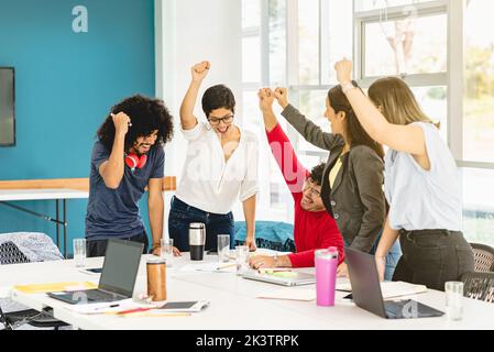Group of excited Hispanic colleagues celebrating achievement of successful project with fists up while gathering at table in coworking space in Costa Stock Photo
