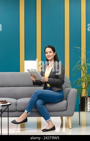 Smiling Hispanic female worker browsing tablet while sitting on sofa in lounge of workspace and thinking about new project in Costa Rica Stock Photo