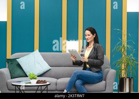 Smiling Hispanic female worker browsing tablet while sitting on sofa in lounge of workspace and thinking about new project in Costa Rica Stock Photo
