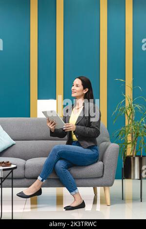 Smiling Hispanic female worker browsing tablet while sitting on sofa in lounge of workspace and thinking about new project in Costa Rica Stock Photo