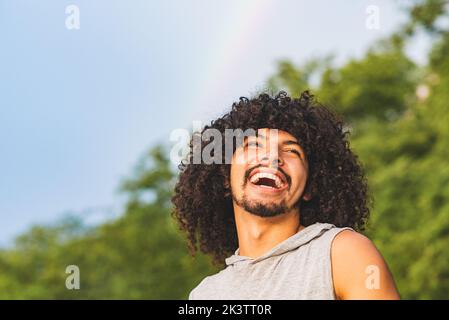 Low angle of laughing young Latin American bearded guy with long curly hair having fun in summer day against green trees and sky with rainbow Stock Photo
