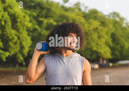 Happy young Latin American guy with long curly hair and beard smiling and looking away while listening to music with portable speaker and spending sum Stock Photo