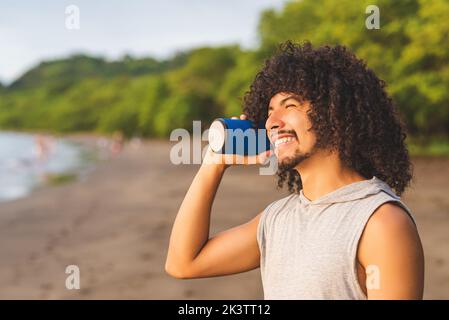 Happy young Latin American guy with long curly hair and beard smiling and looking away while listening to music with portable speaker and spending sum Stock Photo