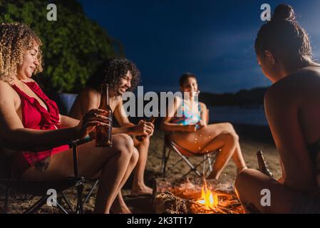 Group of cheerful ethnic friends with beer having fun while gathering around burning bonfire during summer hangout in evening time on beach in Costa R Stock Photo