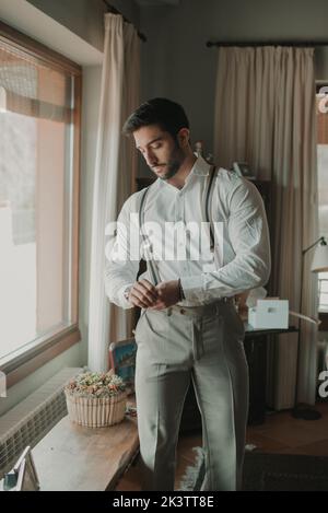 Handsome bearded groom in shirt and trousers with suspenders near seat with composition of blooms and window in room Stock Photo