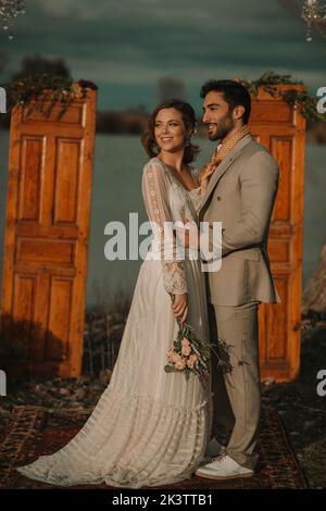 Side view of young guy in grey costume hugging attractive smiling lady in wedding dress and bunch of flowers near decorations in cloudy weather Stock Photo