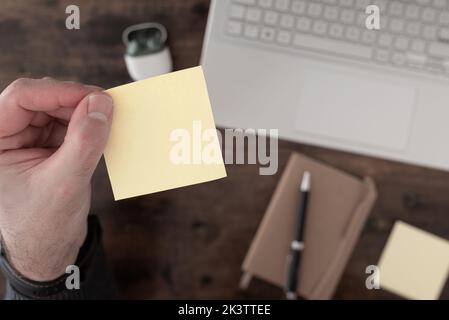 top down view of hand holding empty yellow sticky note above desk with laptop computer and notepad Stock Photo