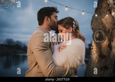 Side view of young guy in grey costume hugging and kissing attractive smiling lady in wedding dress Stock Photo