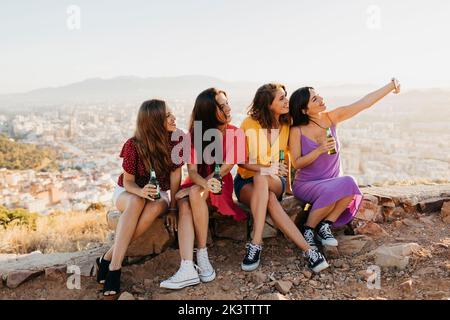 Cheerful group of young female friends in casual outfits taking selfie on smartphone while sitting on rocky ground on hilltop and drinking beer Stock Photo