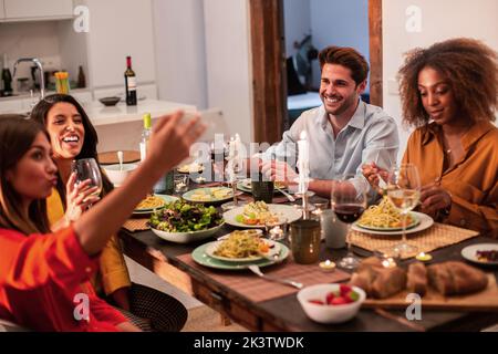 From above happy young man sitting next to black woman and looking at female friend taking selfie on smartphone while having dinner together at home Stock Photo