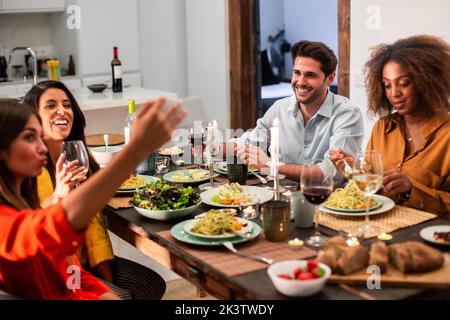 From above happy young man sitting next to black woman and looking at female friend taking selfie on smartphone while having dinner together at home Stock Photo