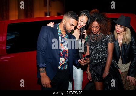 Crop smiling multiracial friends talking and watching cellphone near transport during party in night town Stock Photo