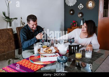 Cheerful young Latin American married couple in casual clothes smiling while having breakfast together at home Stock Photo