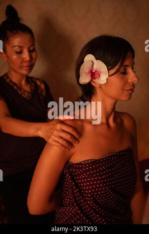 Asian female massagist in traditional clothes giving a thai massage to a caucasian woman sitting on a bed in a room lit with warm lights Stock Photo