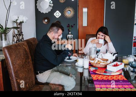 Cheerful young Latin American married couple in casual clothes smiling while having breakfast together at home Stock Photo