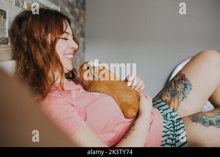 side view of young woman in pajamas playing with Guinea pig and relaxing on bed in bedroom Stock Photo